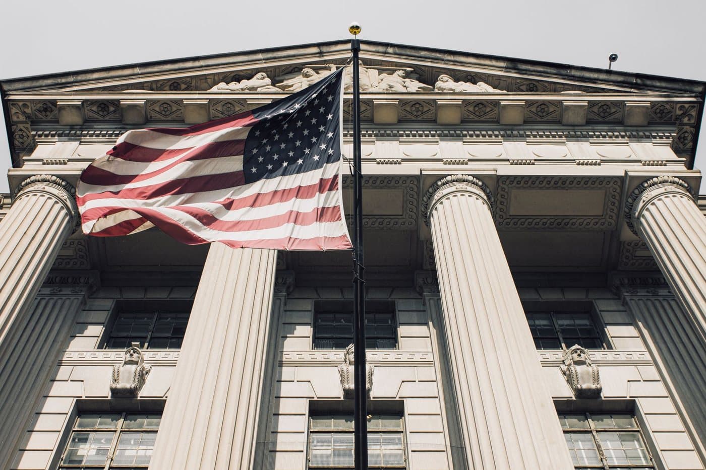A United States flag flying in front of the columned stone facade of a neoclassical federal government building