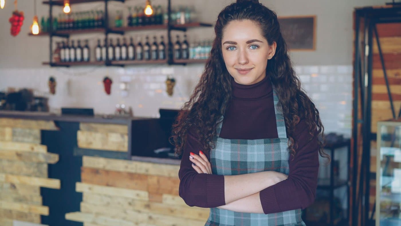A cafe owner in a checked apron standing with arms folded in front of the counter of a small, warmly lit shop