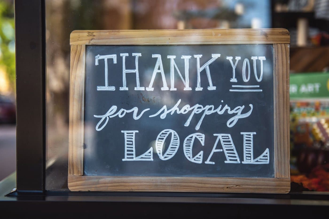 A chalkboard sign in a shop window reading Thank you for shopping local in white hand lettering