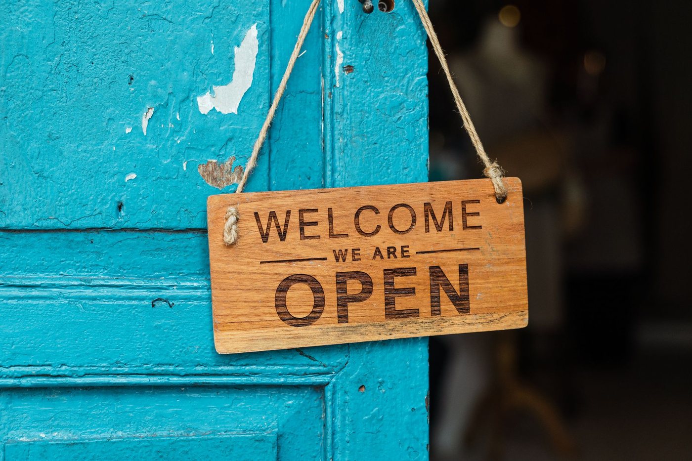 A wooden sign reading Welcome We Are Open hanging by twine on a weathered turquoise painted door