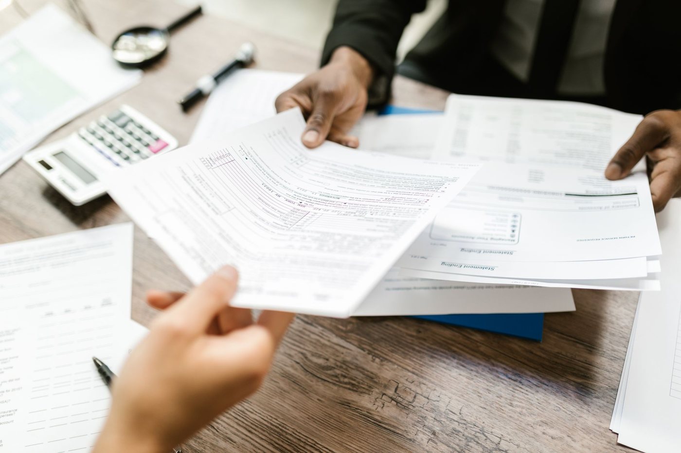 Two people at a desk passing printed financial forms between them, with a calculator and a magnifying glass on the wooden surface
