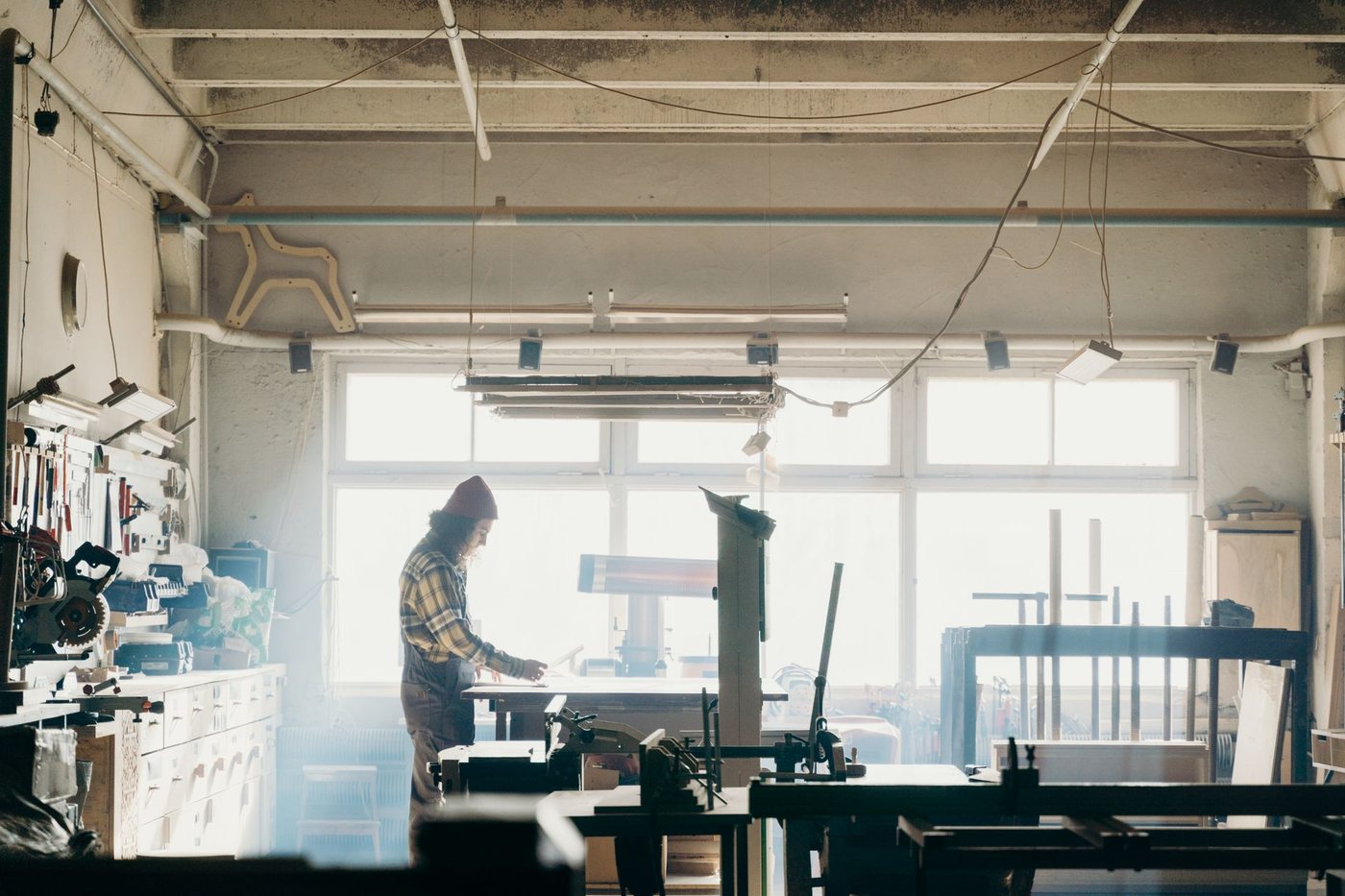 A lone woodworker in a beanie and plaid shirt standing at a bench in a bright, tool-filled workshop with large windows behind