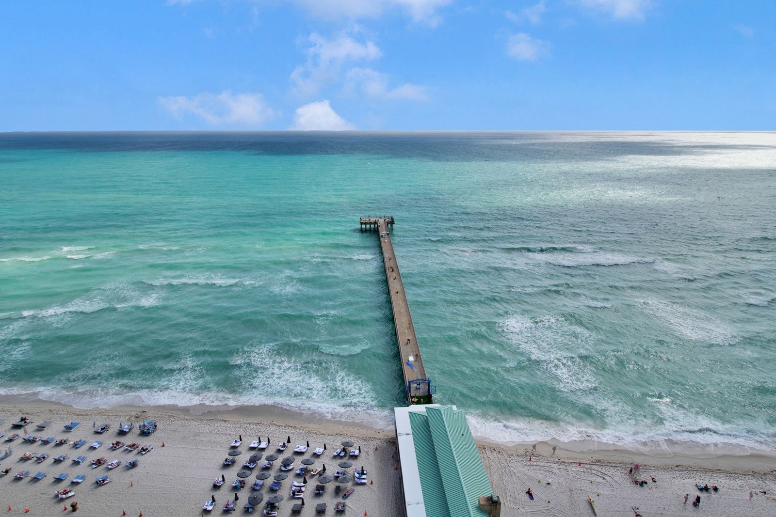 Aerial view of a fishing pier reaching into turquoise water beside a wide beach